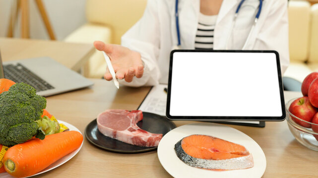 Young Asian female nutritionist in a white lab coat, working at a desk. She shows tablet with blank screen consultation details, promoting weight loss and balanced diets
