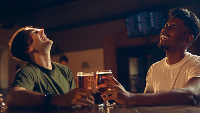 Friends laughing together while holding beer glasses at bar table. Concept of nightlife imagery, bar lifestyle promotion, brewery marketing, beverage advertising.