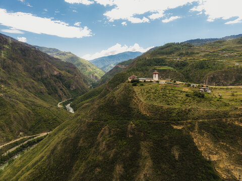 Aerial view of a striking white building with red accents perched atop a hill overlooking a river valley amidst rolling green mountains, Raga, Paro, Bhutan.