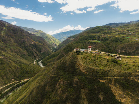 Aerial view of a temple complex perched on a verdant hill overlooking a winding river, the sky a canvas of azure and wispy clouds, Raga, Paro, Bhutan.