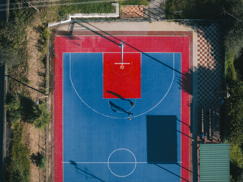 Aerial view of a vibrant basketball court, a canvas of red and blue hues under the sun's gaze, bordered by lush greenery, Thimphu, Bhutan.