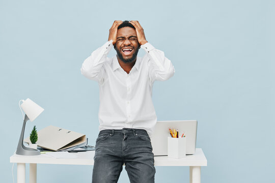 Confused man in white shirt holding head, standing behind modern office desk with open notebook, potted plant, and stationery, expressing frustration or stress, isolated on light blue background