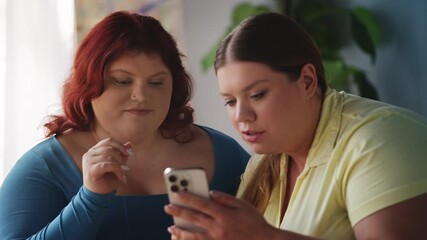 Two happy overweight women scrolling social media on phone, enjoying fun time