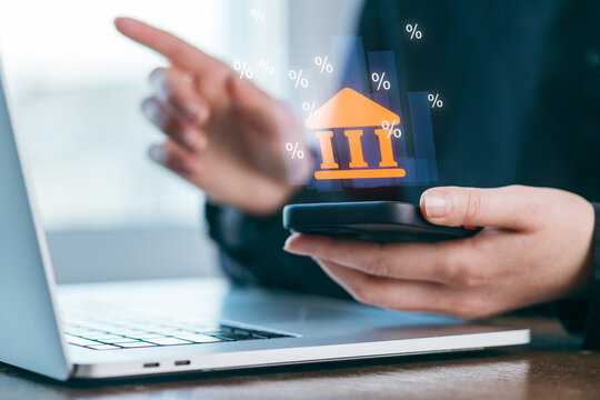 Person using smartphone to manage banking tasks while sitting at a desk with a laptop during daytime