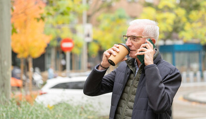 Side view of senior man multitasking, drinking coffee and having a phone call on a city street with copy space