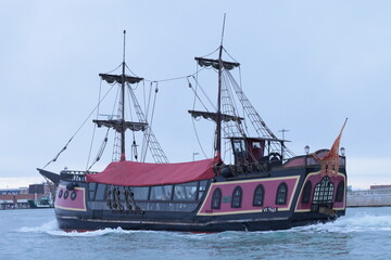 Colorful pirate-style ship with red canopy sailing on the Venetian lagoon near Venice, Italy. The vintage vessel moves across calm water under a cloudy sky.