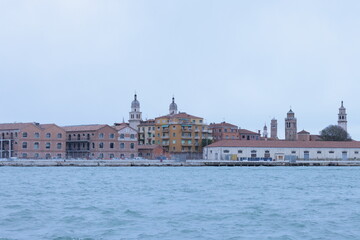 Venice waterfront view with historic brick buildings, church domes, and bell towers under a cloudy sky. Calm lagoon water reflects the charming Italian architecture.