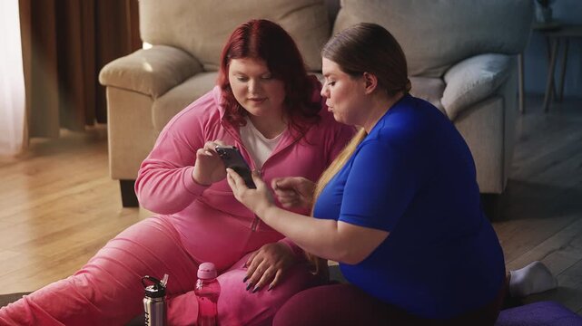 Two curvy women scrolling on phone using fitness tracking app, planning workout