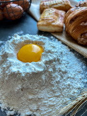 Vertical high-angle shot of a raw egg yolk inside a mound of white flour, surrounded by fresh puff pastries and a wire basket of brown eggs on a dark rustic surface.