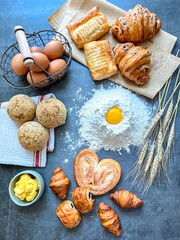 Top-down flat lay composition of a bakery workspace featuring a central mound of flour with a raw egg yolk, surrounded by fresh croissants, sesame buns, puff pastries, a basket of brown eggs, butter, 