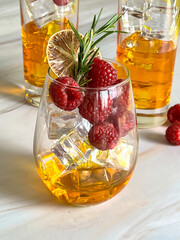 Close-up of a sparkling golden cocktail garnished with raspberries, rosemary, and dried lemon in a stemless glass, served on a white marble table with blurred drinks in the background.