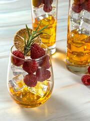 Close-up of a sparkling golden cocktail garnished with raspberries, rosemary, and dried lemon in a stemless glass, served on a white marble table with blurred drinks in the background.