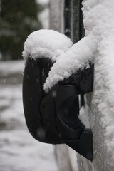 Der Seitenspiegel eines wei&szlig;en Autos ist vollst&auml;ndig mit Schnee bedeckt, von hinten auf einem vertikalen Foto sichtbar. Die Nahaufnahme betont die Schneetextur und winterliche Autoatmosph&auml;re.