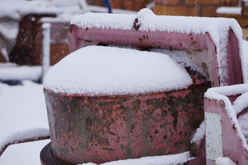 Stara czerwona betoniarka pokryta śniegiem stoi na placu budowy. W tle widać domy i białe zaspy, zimowa sceneria budowlana. © Adam