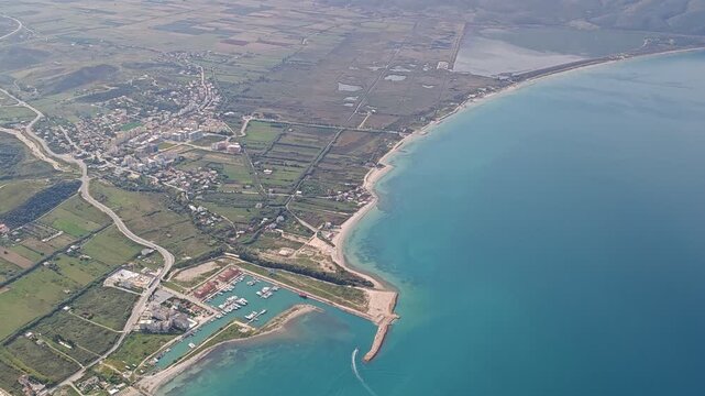 Aerial panorama of Orikum, Albania, showcasing the seaside town, turquoise coastline, marina, and surrounding mountains under clear daylight for a vibrant Mediterranean view.