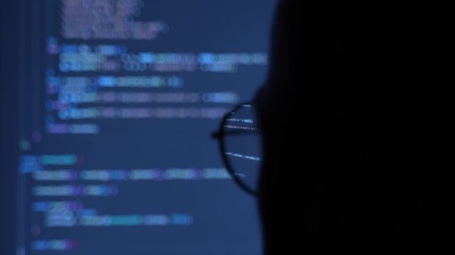 Programmer silhouette working on a computer at night with code reflecting on glasses