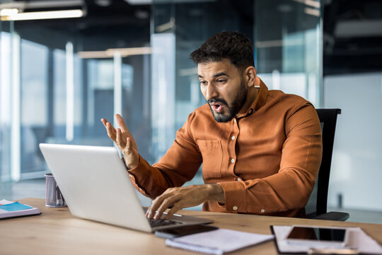 Businessman looking surprised and stressed while receiving bad news or an unexpected problem on his laptop, expressing astonishment with open mouth and spread hand gesture in a modern office setup