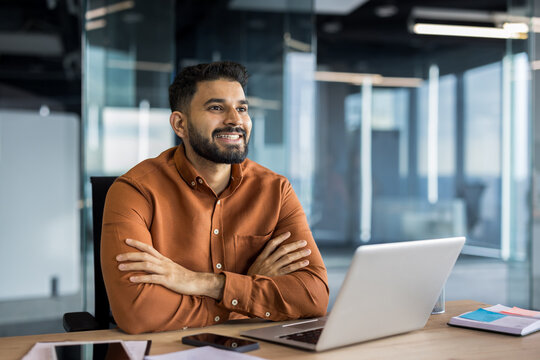 Smiling indian businessman in a brown shirt sitting with crossed arms at an office desk, looking away with a confident expression, using a laptop and focusing on future business success