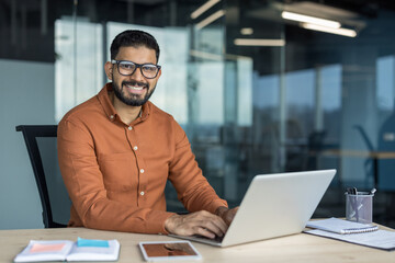 Indian businessman sitting at his desk, typing on a laptop and smiling confidently, representing success, productivity, and a positive professional attitude in a contemporary office environment