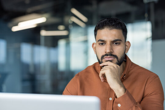 Young bearded man feeling pensive and concentrated, holding chin while looking at a laptop screen in a modern office, deep in thought, brainstorming new ideas for business
