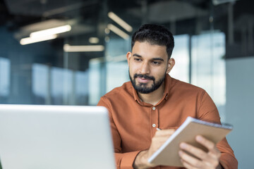Young man concentrating on his work, looking at a laptop screen and writing in a notebook in a...