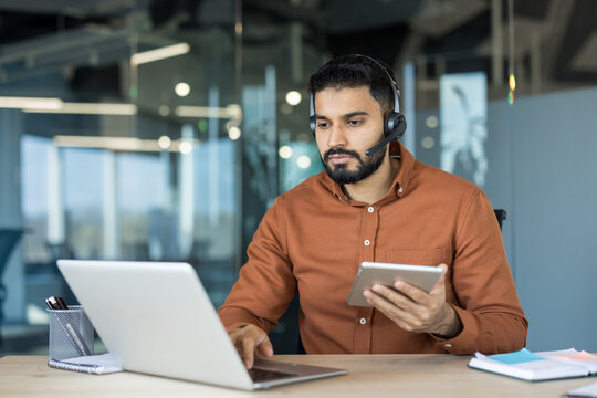 Young male contact center agent wearing a headset and using a laptop with a digital tablet, providing support and communication in a professional office environment