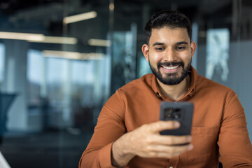 Young indian businessman smiling and looking at his mobile phone while connecting online from a modern blurred office, conveying remote work, networking, and digital productivity