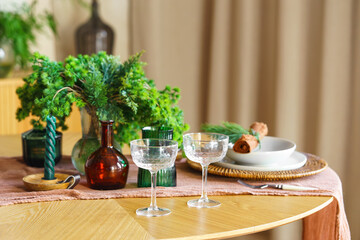 Close up of elegant crystal glasses, plates and flowers on wooden table with green decorations and warm holiday ambiance. Perfect for celebrations, cozy and romantic gatherings, seasonal home decor.