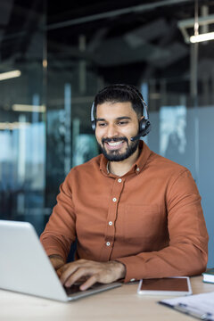 Smiling indian customer service agent wearing headset, typing on laptop at modern office desk while providing online support and friendly it helpdesk assistance to clients remotely