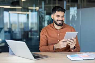 Smiling young professional man with a beard sitting at his desk, cheerfully using a digital tablet while a laptop and planner are on the table, indicating a modern and efficient work environment