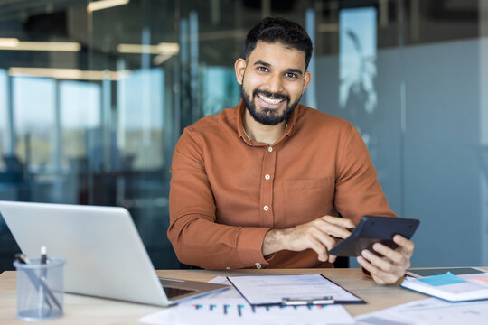 Smiling indian businessman calculating expenses at a modern office desk with laptop, calculator and documents, analyzing financial data for accurate accounting and successful management - Powered by Adobe