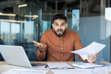 Young businessman sitting at his desk with a laptop and documents, expressing confusion and stress with wide eyes and open mouth, working in a modern office environment