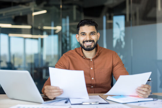 Indian businessman sitting at a modern office desk, smiling at the camera while holding and reviewing important documents, reflecting productivity and success in a corporate environment