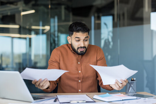 Confused businessman holding two documents, comparing data and reports while feeling stress and uncertainty about financial information and paperwork in office