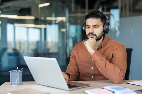 Young man wearing headphones thoughtfully working on a laptop in a modern office, engaged in focused online learning or virtual meeting while observing the screen
