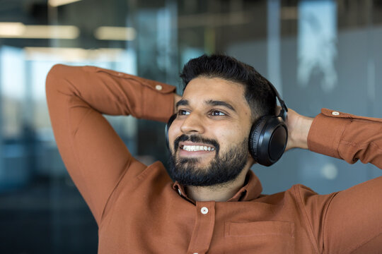 Young man wearing headphones and dark brown shirt, smiling with hands behind his head, relaxing and enjoying break time while looking away in bright indoor setting
