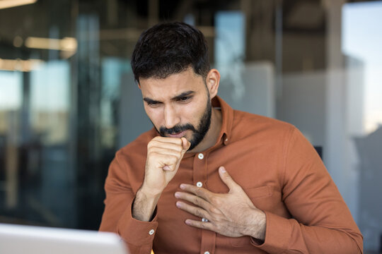 Young man experiencing an illness, coughing and clutching his chest, indicating discomfort and potential respiratory issues while trying to work in a modern office environment
