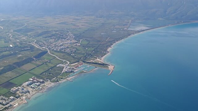 Aerial panorama of Orikum, Albania, showcasing the seaside town, turquoise coastline, marina, and surrounding mountains under clear daylight for a vibrant Mediterranean view.