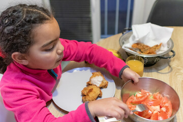 Young girl enjoying a home cooked meal, sitting at the table, learning healthy eating habits, balancing fried chicken with fresh tomato salad, showing childhood food choices