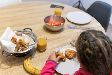 Young girl with braided hair eating crispy fried chicken with her hands at a wooden dining table, enjoying a casual homemade meal with fresh juice and salad