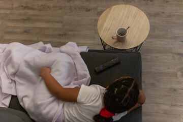 Young girl relaxing and watching tv on a comfortable couch at home, enjoying leisure time and entertainment while being cozy under a blanket