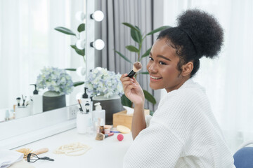 Joyful african teenage girl holding makeup brush while looking sideways smiling in beauty routine wearing robe seated at vanity table with cosmetics, flowers, mirror, and green indoor plants