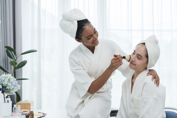 african american teenage girl using makeup brush on asian teenage friend both wearing white robes smiling during relaxing beauty session skincare and friendship lifestyle selfcare concept confidence