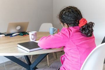 Young girl with braided hair and pink jacket busy concentrating on her schoolwork at a wooden table, diligently writing in her notebook during a study session