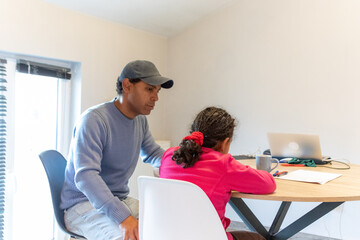 Dad is supporting his young daughter with her schoolwork, patiently guiding her through a task at a wooden table with a laptop and paper, illustrating family learning and education