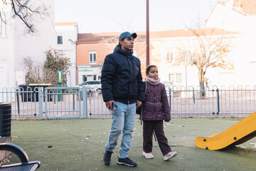 Father and daughter walking hand in hand through an urban park playground on a dry day, representing family bonding, care, protection, and shared moments