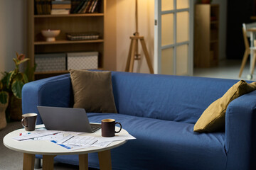 Modern living room featuring open laptop, coffee mugs, scattered tax documents and financial papers on round table, suggesting remote work or individual preparing taxes at home