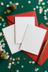 White blank stationery sheets arranged on a green surface, surrounded by colorful confetti and red envelopes, creating a festive atmosphere for invitations or greeting cards