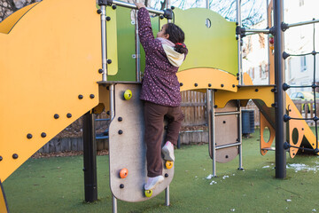 Young girl actively climbing playground structure, reaching for metal bar, playing outdoors and developing physical skills on colorful equipment during winter day