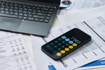 Smartphone displaying calculator app lying on financial documents near laptop, calculating tax payments and analyzing expenses during tax season, close up of modern workspace